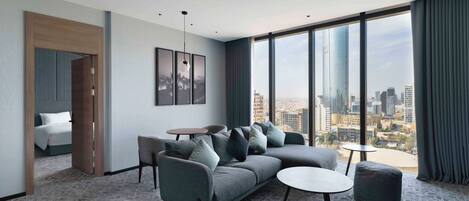 A modern hotel room with a large window offering a city view, a grey sofa, a round coffee table, and a small ottoman.
