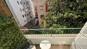 Ein Balkon mit einem grünen Stuhl, einem kleinen Tisch und Blick auf benachbarte Gebäude und Grünflächen.