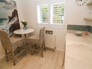 A room with a round table, two wicker chairs, a vase with flowers, a clock, and a window with blinds. - Sandwaves (St Ives)