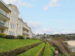 A row of multi-story residential buildings with balconies, a grassy lawn, and a pathway leading to a beach. - Sandwaves (St Ives)