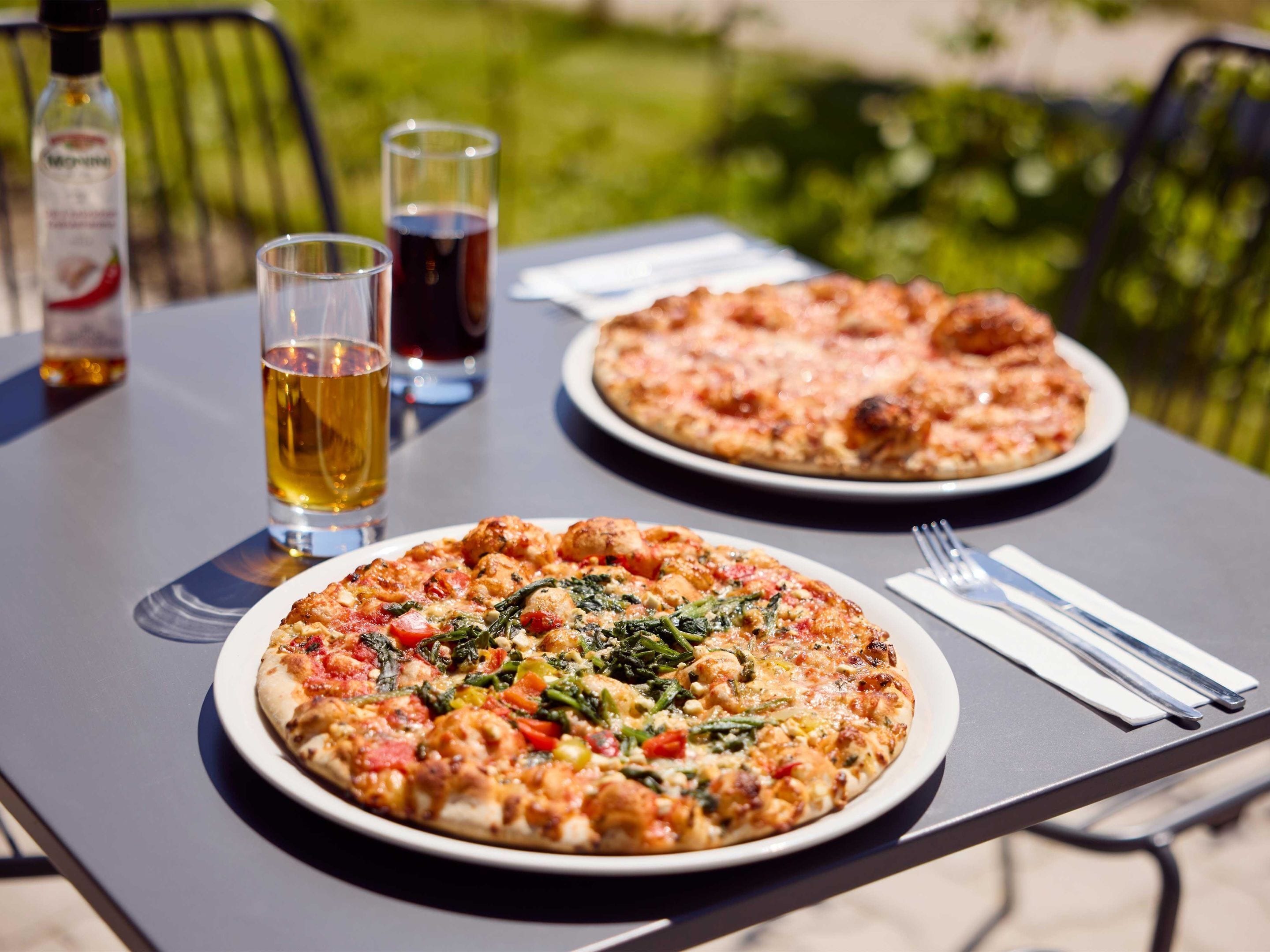 Dos pizzas en platos, con una botella y vasos sobre una mesa al aire libre.