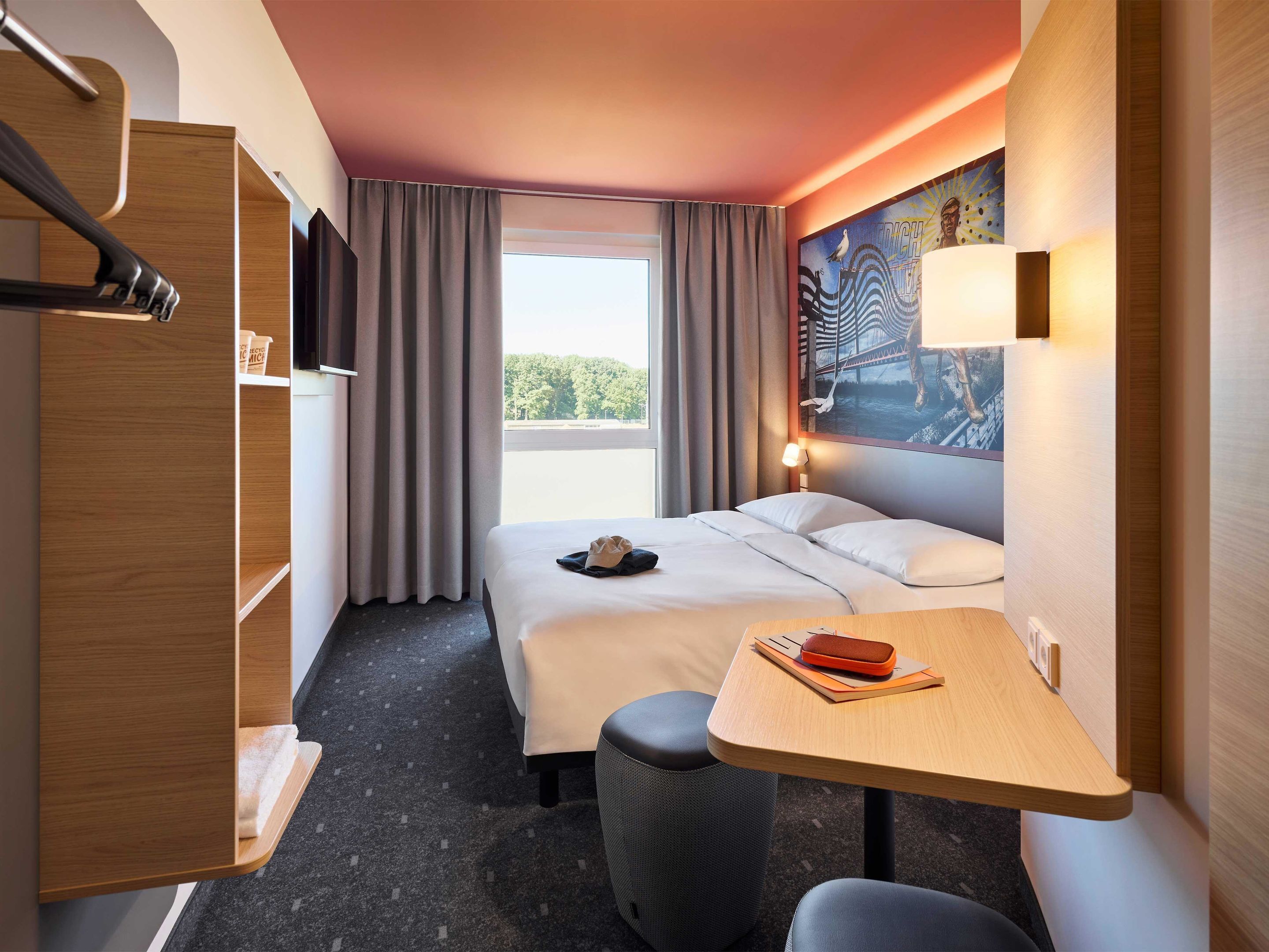 Habitación de hotel con cama, escritorio con lámpara, silla, armario y ventana grande con cortinas.