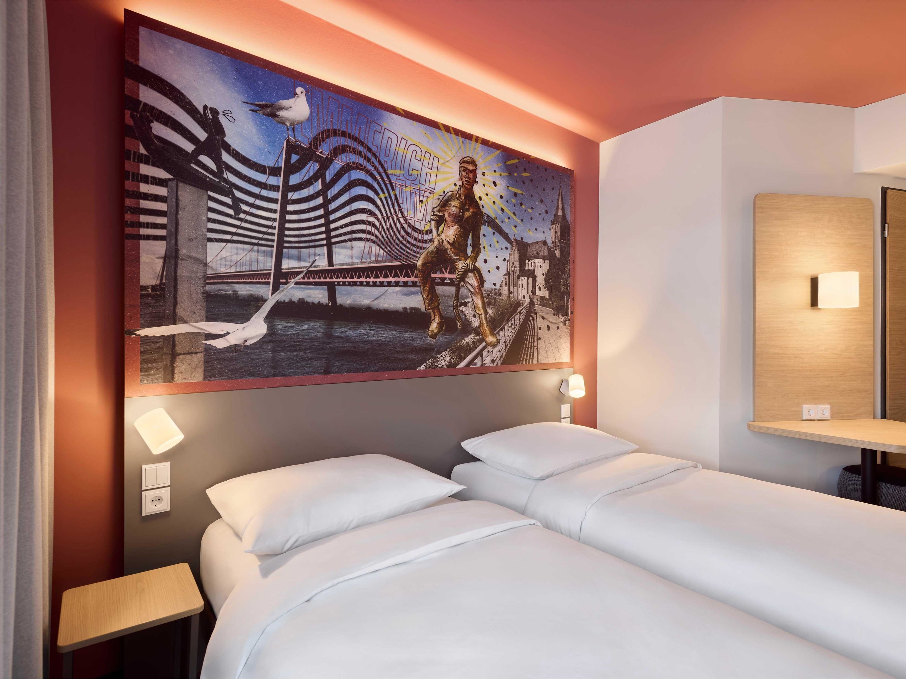 A hotel room with a large mural on the wall, a bed with white bedding, a small bedside table, and a wall-mounted light fixture.