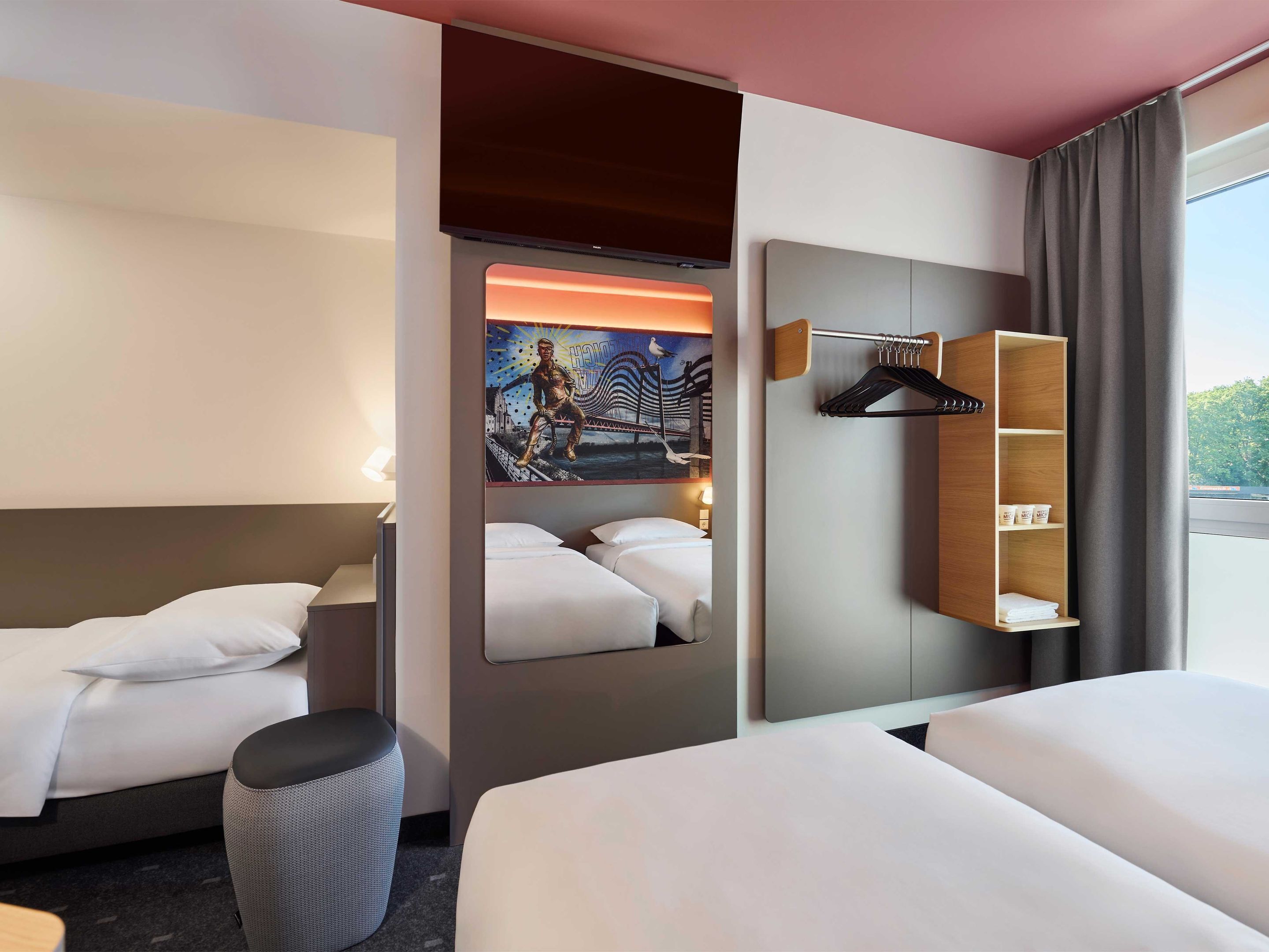 A hotel room with two beds, a large mirror, a painting, and a wooden shelf with toiletries.