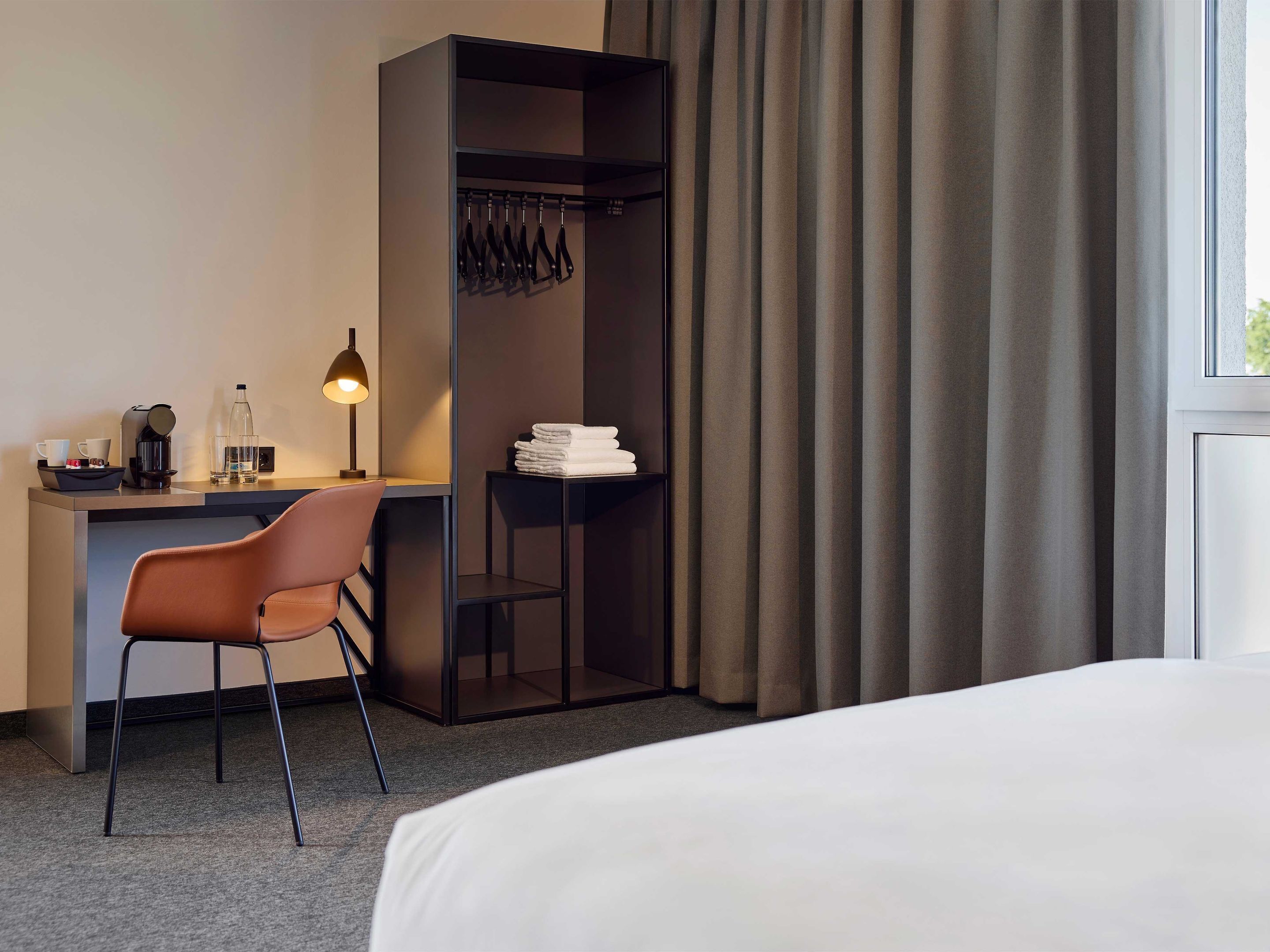 Habitación de hotel con cama, escritorio con silla, lámpara, perchero y ventana con cortinas.