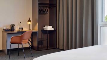 Habitación de hotel con cama, escritorio con silla, lámpara, perchero y ventana con cortinas.