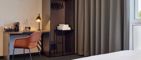 A hotel room with a bed, a desk with a chair, a lamp, a coat rack, and a window with curtains.