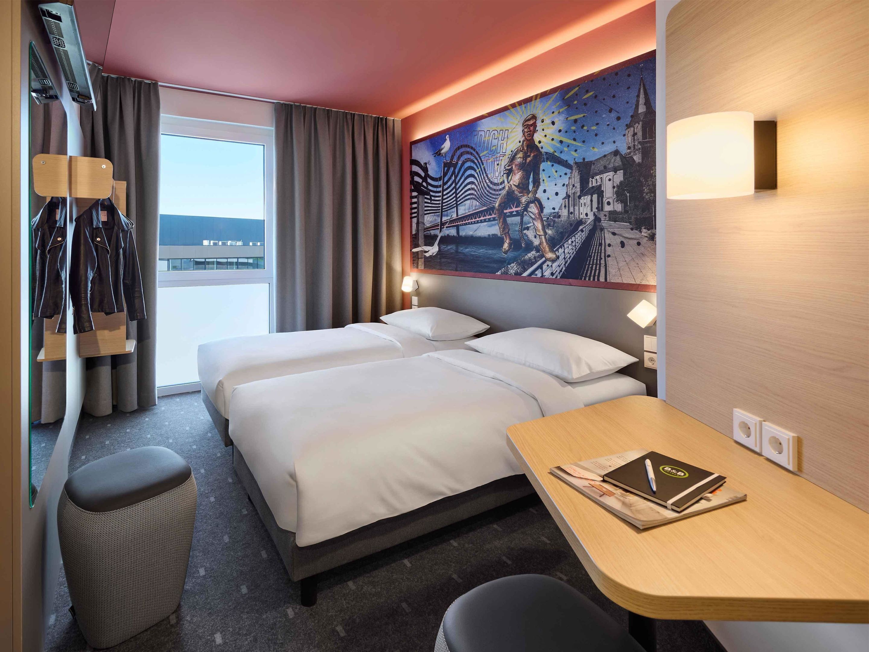 Une chambre d’hôtel avec deux lits, un bureau avec un ordinateur portable, un porte-manteau et une grande fresque murale.