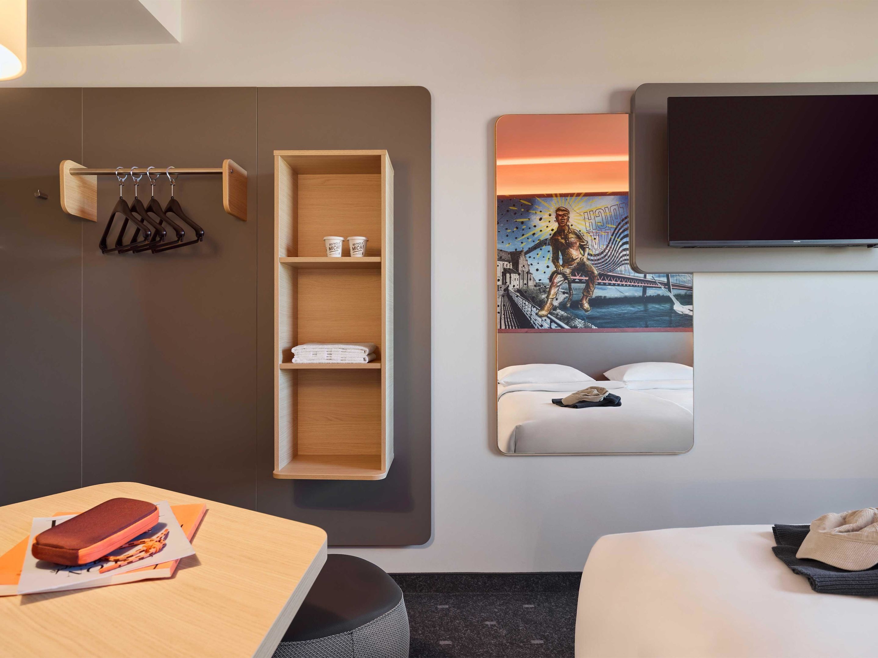 Une chambre d’hôtel avec une étagère en bois, un lit avec une literie blanche, un téléviseur à écran plat fixé au mur et une table sur laquelle se trouvent un sandwich et des serviettes en papier.