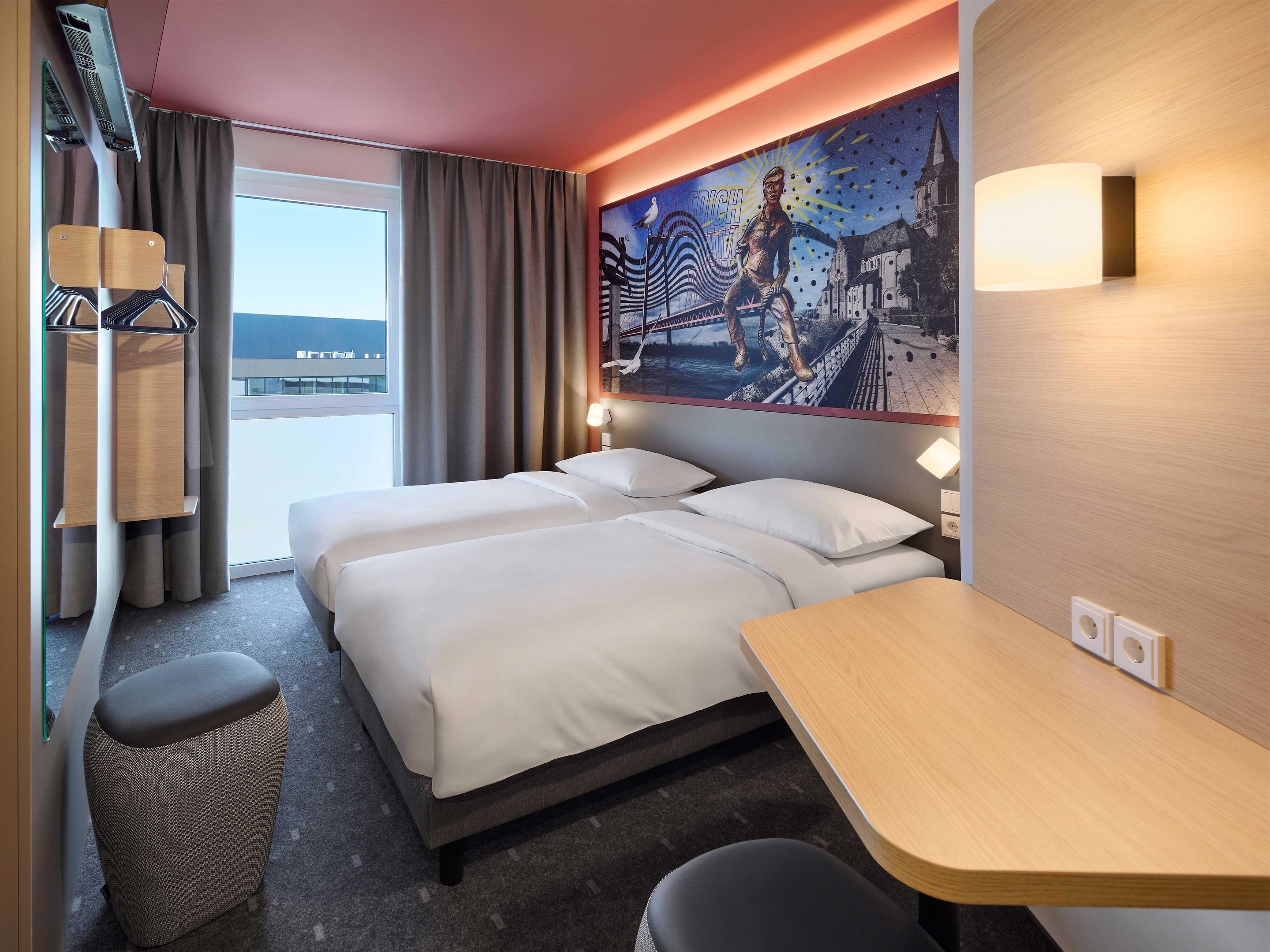 Habitación de hotel con dos camas, un escritorio pequeño, una silla, una ventana con cortinas, un mural en la pared y un ventilador de techo.