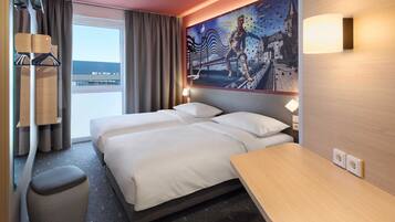 Habitación de hotel con dos camas, un escritorio pequeño, una silla, una ventana con cortinas, un mural en la pared y un ventilador de techo.