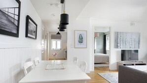 Un comedor moderno y luminoso con una mesa blanca, sillas y un florero con flores. Se ve una cocina al fondo con un televisor y un sofá.