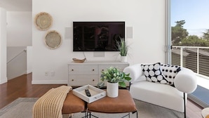 Una sala de estar moderna con un televisor de pantalla plana montado en la pared, un sofá blanco con cojines estampados, una mesa de centro con un control remoto y una planta en maceta.