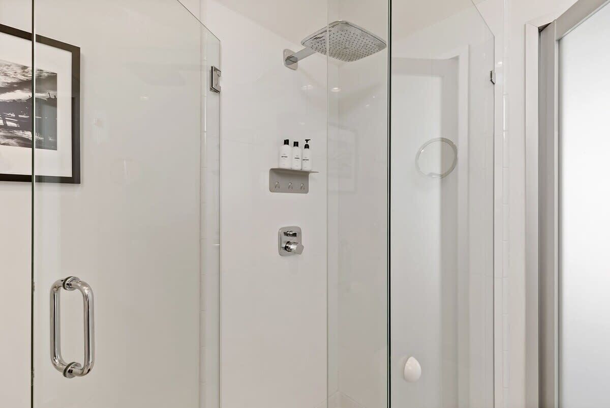 A modern shower with a glass door, a mounted shelf with toiletries, and a framed picture on the wall.