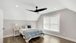Ein Schlafzimmer mit einem Bett, einem Nachttisch, einem Deckenventilator, einem Fenster mit Jalousien und Holzböden.