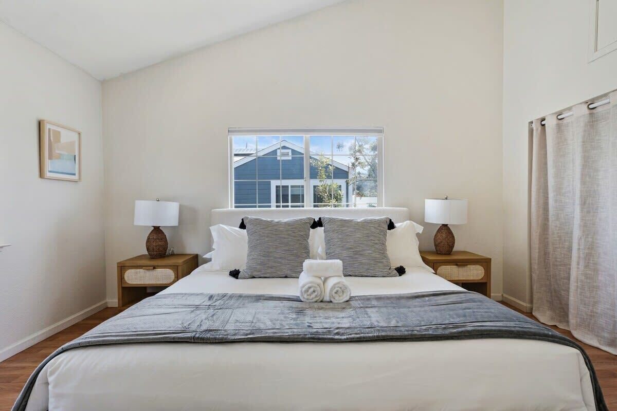 A bedroom with a large bed, two bedside tables with lamps, a window with a view of a house, and a curtain on the right.