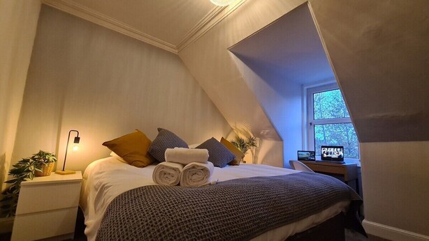 A bedroom with a sloped ceiling, a bed with rolled towels, pillows, and a blanket. There is a bedside table with a lamp and a plant. A laptop is on a desk next to the bed. - 5 Bed Sleeps 10 Full Kitchen Parking (South Ayrshire Council)