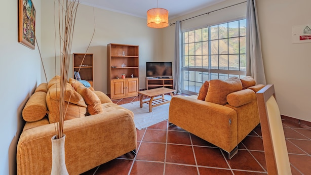 A living room with two orange sofas, a wooden cabinet, a TV, and a large window with a view of the outdoors. - Simões Apartment by HR Madeira (Porto Santo)