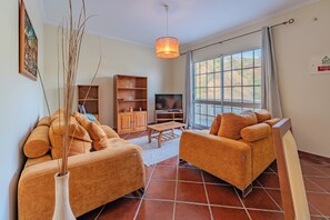 A living room with two orange sofas, a wooden cabinet, a TV, and a large window with a view of the outdoors. - Simões Apartment by HR Madeira (Porto Santo)