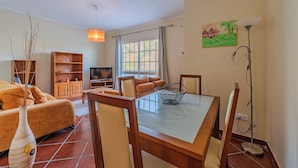 A living room with a glass-top dining table, wooden chairs, a sofa, a TV, and a bookshelf. - Simões Apartment by HR Madeira (Porto Santo)