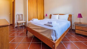 A neatly made bed with white linens and a folded towel, a wooden nightstand with a lamp, and a chair in a room with tiled flooring and wooden walls. - Simões Apartment by HR Madeira (Porto Santo)
