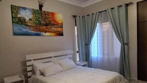 A bedroom with a white bed, a nightstand, a painting on the wall, and light blue curtains. - BLT Apartments - classy (Kira Town)