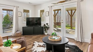 Ein modernes Wohnzimmer mit einem großen Flachbildfernseher, Holzboden, einem runden Couchtisch und Blick auf eine Holzterrasse und Bäume durch Glastüren.