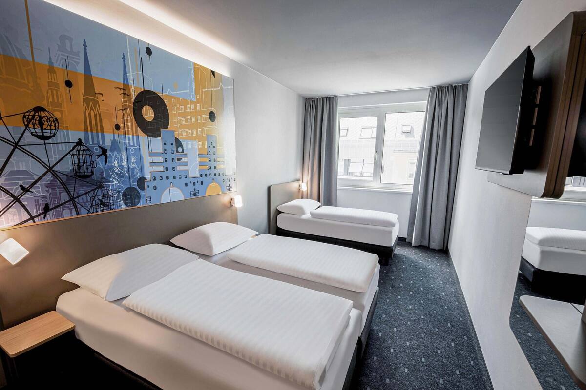 Une chambre d’hôtel avec deux lits, un bureau, une chaise, une télévision et une grande fenêtre avec des rideaux.