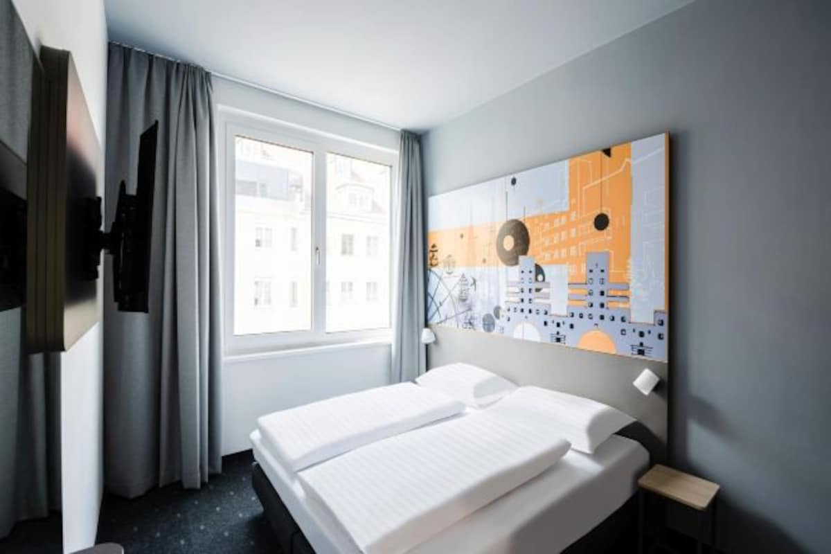 A hotel room with a bed, a window with curtains, a painting on the wall, and a nightstand.