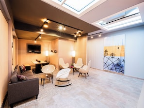 A modern living room with a high ceiling, skylights, a flat-screen TV, a dining area, and a seating arrangement with a rocking chair and armchairs. - B&B HOTEL Wien-Heilligenstadt (Vienna)