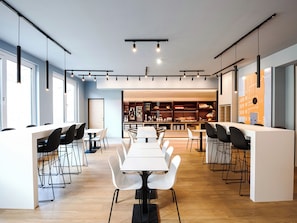 A modern dining area with a long table and chairs, a bar area with shelves of beverages, and a view of the cityscape through large windows. - B&B HOTEL Wien-Heilligenstadt (Vienna)