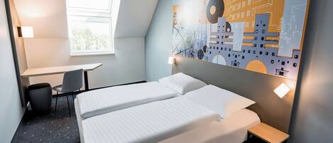 A hotel room with two beds, a desk, a chair, a window, and a wall with a cityscape mural.