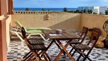 Une terrasse avec un sol à carreaux, une table et des chaises en bois, un coussin vert, et une vue sur la mer et les bâtiments en arrière-plan.