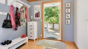 Een kamer met een houten plafond, een witte bank met schoenen, een kapstok met kleding, een dressoir en een deur die uitkomt op een tuin.
