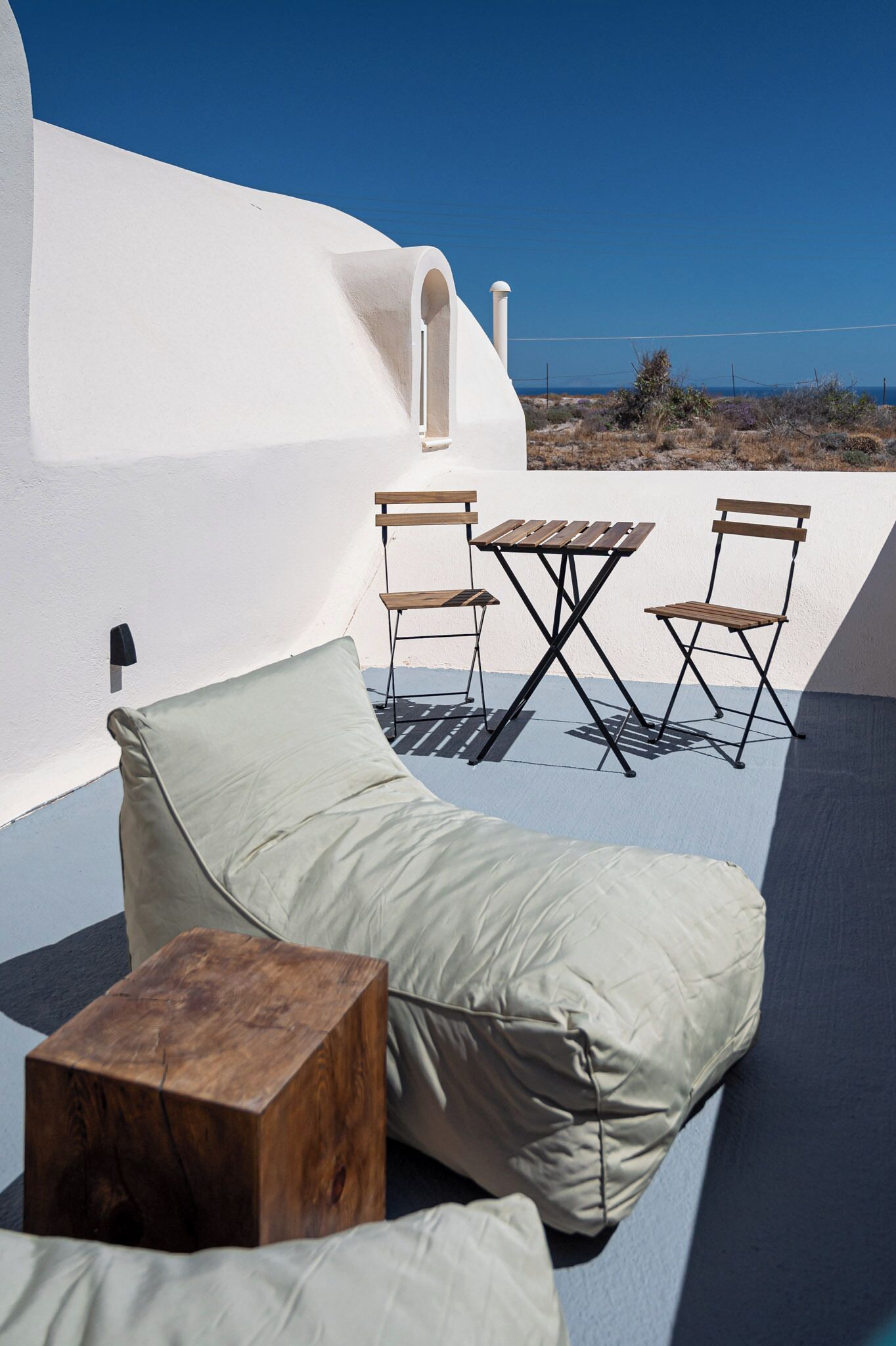 Un área de estar al aire libre iluminada por el sol, con una silla lounge acolchada, un taburete de madera y una mesita.