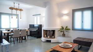 A spacious living room with a fireplace, a piano, and a dining area with chairs and a table. - Villa Chora (Santorini)