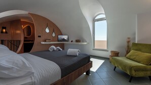 A modern bedroom with a curved ceiling, a bed with white and dark bedding, a green armchair, and a small shelf with decorative items. - Villa Chora (Santorini)