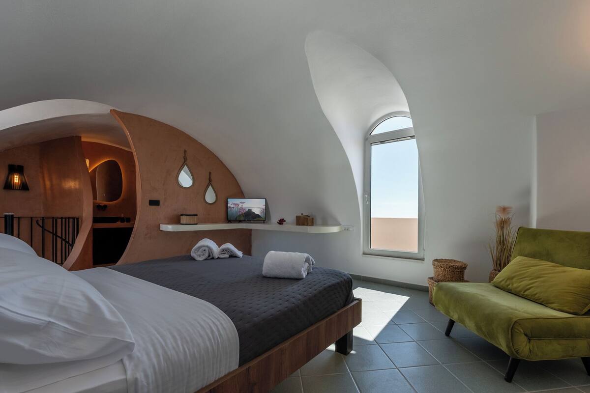 A modern bedroom with a curved ceiling, a bed with white and dark bedding, a green armchair, and a small shelf with decorative items.