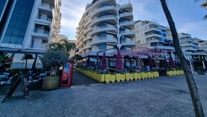 Un immeuble à plusieurs étages doté de balcons, d’un distributeur automatique de billets rouge et d’un espace extérieur aménagé avec des parasols.