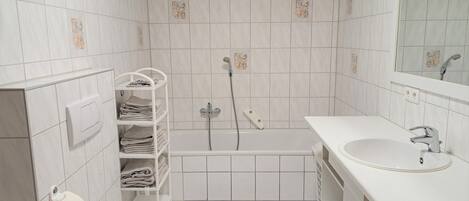 Ein kleines, sauberes Badezimmer mit weißen Fliesen, einer Toilette, einem Waschbecken, einer Dusche und einem Handtuchhalter.