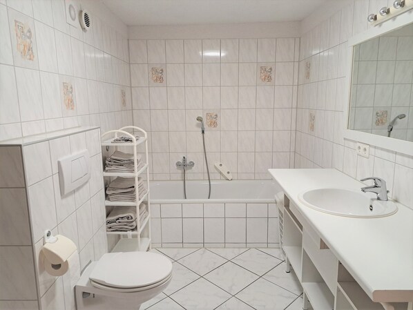 Un baño pequeño y limpio con azulejos blancos, inodoro, lavabo, ducha y un perchero para toallas.