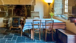 A rustic interior with a stone fireplace, wooden beams, a table with chairs, and a vase of flowers. - Métairie du Hellès (Lanmeur)