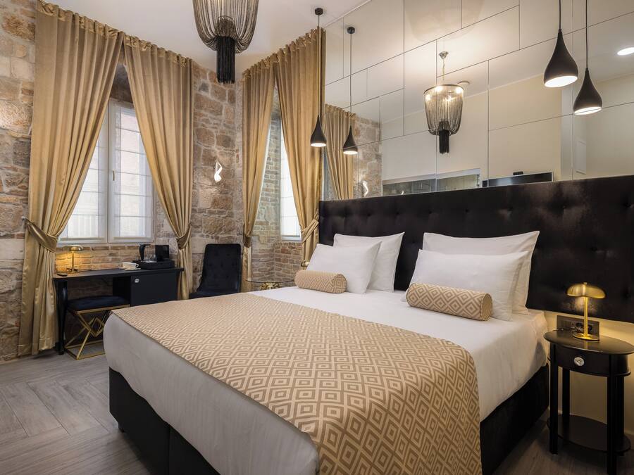 Une chambre d’hôtel moderne avec un grand lit, un bureau et une chaise. La chambre est dotée de murs en pierre, d’une grande fenêtre avec des rideaux et de luminaires suspendus.