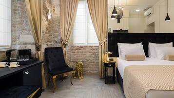 Hotelkamer met een bakstenen muur, een groot bed met witte lakens, een zwart nachtkastje, een gouden beeld van een leeuw, een zwarte fauteuil en een bureau met een koffiezetapparaat en kopjes.