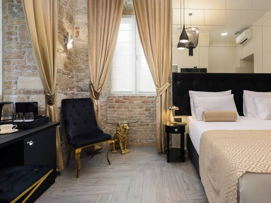 Hotelkamer met een bakstenen muur, een groot bed met witte lakens, een zwart nachtkastje, een gouden beeld van een leeuw, een zwarte fauteuil en een bureau met een koffiezetapparaat en kopjes.
