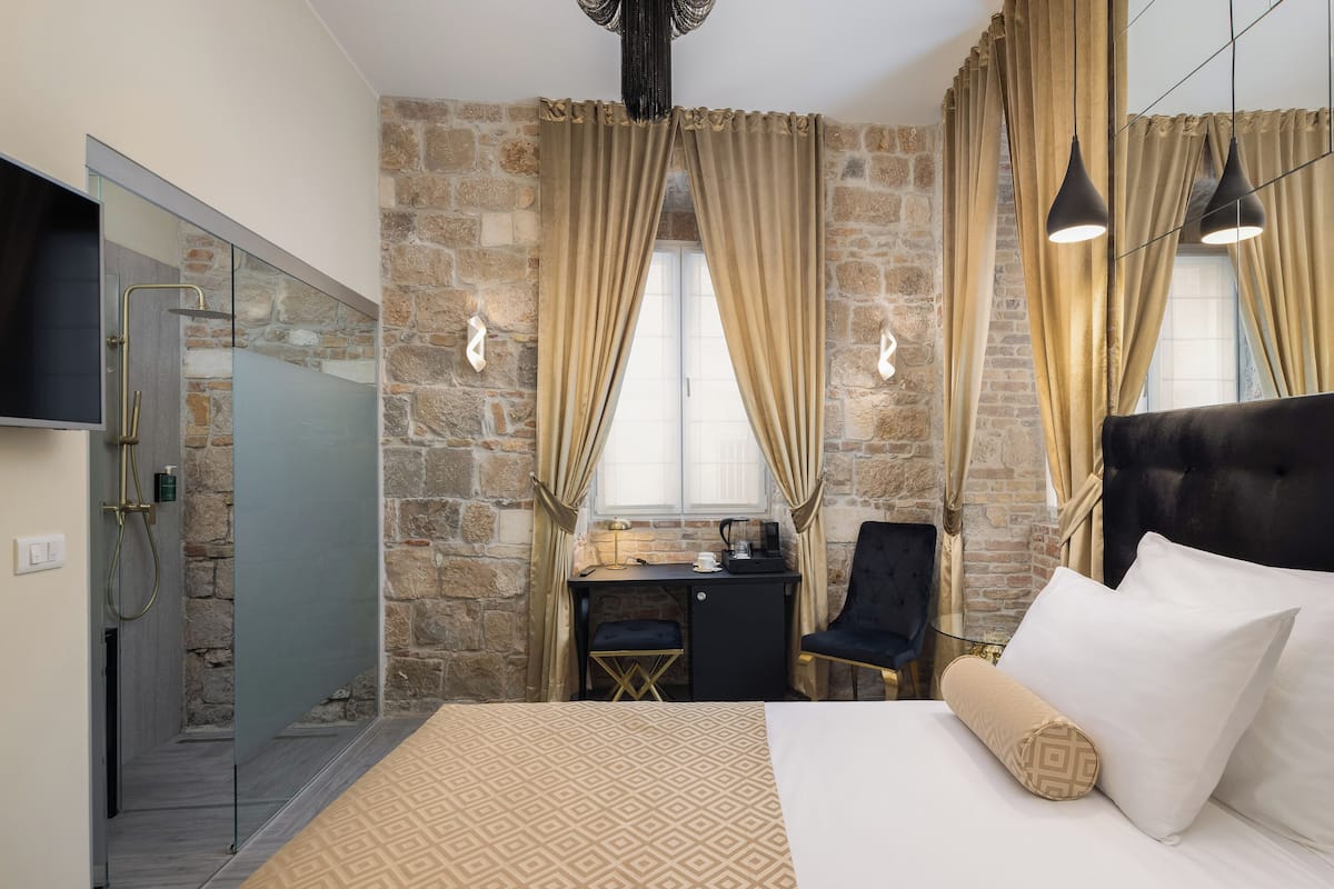 A hotel room with a stone wall, a bed with white and patterned bedding, a desk with a chair, and a shower area.