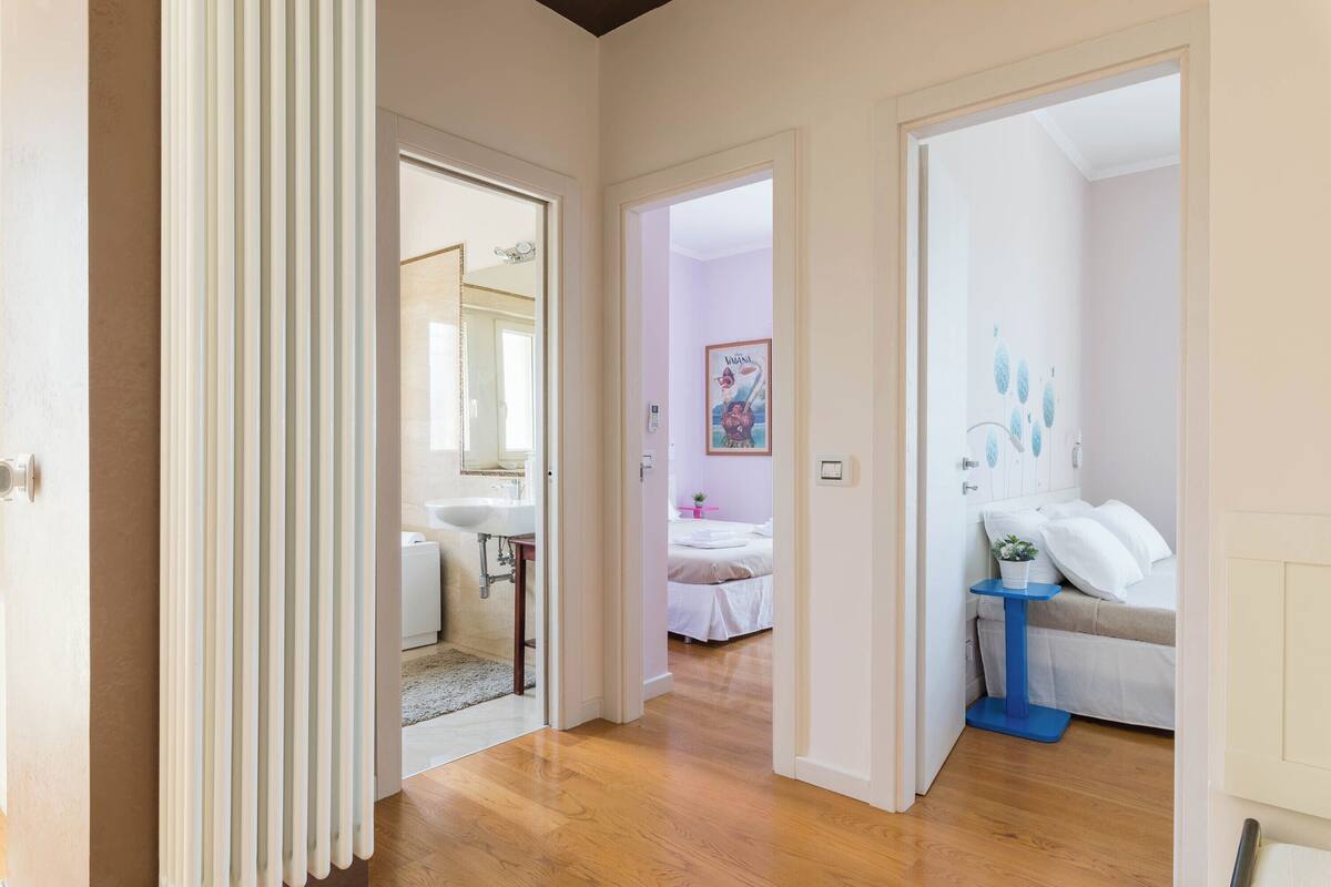 Un corridoio conduce a una camera da letto con un letto, un tavolino e un comodino. Si intravede anche un bagno con lavandino e specchio.