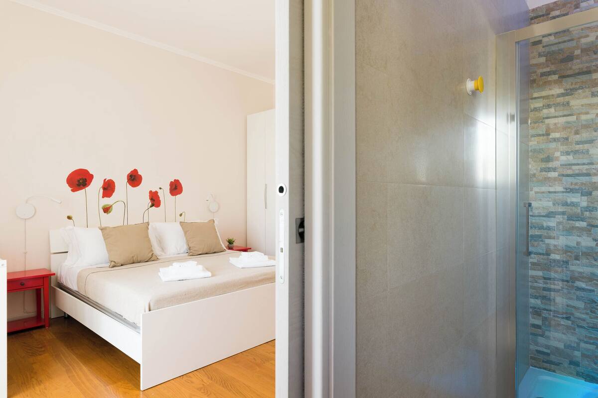 Camera da letto moderna con un letto, un comodino, una lampada rossa e un angolo doccia con porta in vetro.