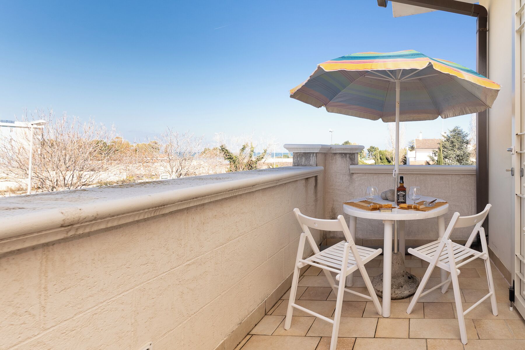 Un balcon avec une table et des chaises, un parasol coloré et une bouteille de spiritueux.