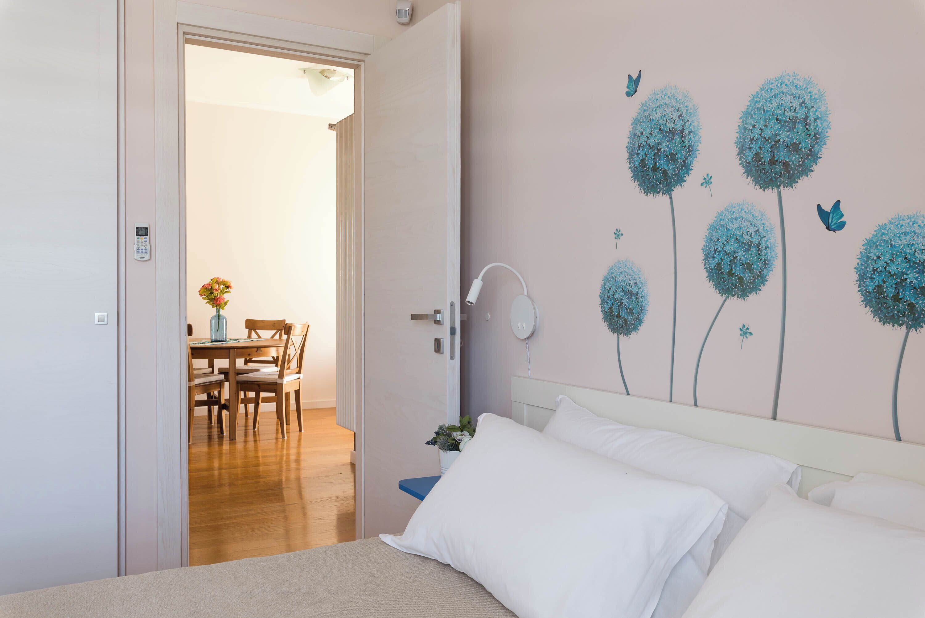 A bedroom with a bed, a wall-mounted lamp, and a dining area visible through an open door.